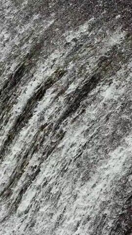 This is a close-up video of a waterfall. The water is flowing rapidly over rocks, creating a foamy and dynamic texture. The lighting is somewhat dim, with highlights on the water's surface. The colors are primarily shades of gray, white, and dark green. The camera is positioned to capture the movement and texture of the water as it cascades down.