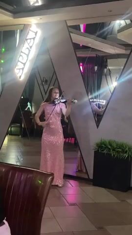 A woman in a sparkling pink floor-length gown plays the violin in what appears to be a restaurant or event space. She is positioned in front of a large, angular mirrored structure that reflects the room. The reflections show tables, chairs, and some decorative elements, including a plant in a dark planter. The lighting is dim with colorful spotlights, casting beams across the scene and the performer. A neon sign with Cyrillic lettering, reading "АРБАЙ" (Arbay), is visible on the mirrored structure. The overall atmosphere suggests a live performance or entertainment during an evening gathering. The woman appears focused on her playing.