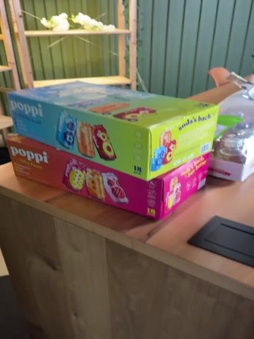 The video shows an indoor scene featuring a wooden table laden with various items. In the foreground, two large boxes of "Poppi" drinks are stacked. The top box is a blend of blue and green, featuring the brand name "Poppi" and the slogan "soda's back, but better," along with images of several fruit flavors and the text "18 pack." Beneath it is a pink and orange box of "Poppi," also indicating an "18 pack" and displaying different fruit flavors.

A person's hand reaches into the frame, touches the top "Poppi" box, and then moves away. To the right of the stacked boxes, other smaller boxes and packages are visible, along with clear plastic bottles or containers and white disposable cups. A black square object rests on the table in the immediate foreground.

In the background, a light green slatted wall can be seen, with a wooden shelf unit positioned against it, holding some white objects. A wooden chair is partially visible in the mid-ground. The person speaking states, "Guys, this is my favorite drink, Poppi. I've got two a day," indicating their fondness for the beverage. The setting appears to be a casual area, possibly a breakroom or an event setup.