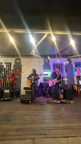 This video shows a live music performance taking place on an outdoor stage at night. Three male musicians are present. The musician on the far left is playing a bass guitar and wearing a red shirt and dark pants. The central musician is playing an acoustic guitar and singing into a microphone, dressed in dark clothing. The musician on the far right is playing an electric guitar and wearing a dark shirt and pants. Behind the musicians, drum equipment is visible, including a drum set. The stage is made of wood, and in the background, there is a building with white doors and windows, illuminated by string lights and spotlights. The overall atmosphere suggests a casual outdoor venue.