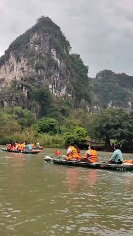 The scene shows several small boats filled with people on a river, likely as part of a tour. The people are wearing bright orange life vests. Each boat has at least one person rowing. There is a large mountain in the background, partially covered in trees. The sky is overcast.