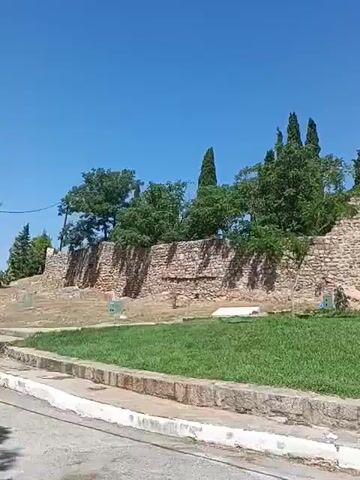 The video shows a historical stone structure with a green lawn in front and trees in the background. The structure appears to be a fortress or castle wall, with an arched entrance visible on the left side. Some people are standing near the entrance. A bell tower with a cross on top is visible on the right side of the structure. The sky is clear and blue. The overall scene suggests a sunny day in a historical site.