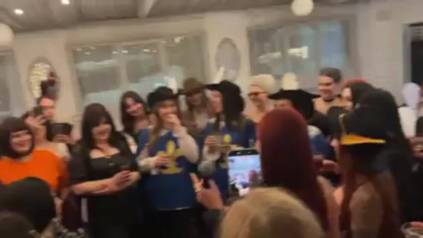 Here is a description of the video:

A group of about 20 women are gathered in what appears to be an indoor event space, possibly a party venue. They are dressed in a variety of costumes, including musketeer outfits, cowboy hats, and other eclectic attire. One woman with red hair holds a microphone and is speaking to the group, while another woman stands in front of her recording with a smartphone. Many of the women are holding glasses filled with a dark liquid, and they raise them in a toast. The atmosphere seems festive and celebratory, with laughter and chatter audible. The background features windows, decorative mirrors, and what looks like drapery or lighting, giving the space a decorated and intimate feel.