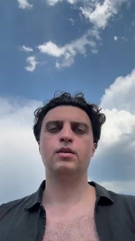 The video begins with a close-up of a man's face against a cloudy sky. The man has dark, curly hair and is wearing a dark open-necked shirt. As the video progresses, the camera pans out to reveal the man standing on a wooden dock. Several boats are visible in the water to his left, and a body of water stretches out behind him. There are white poles with red and white markings along the dock, suggesting it is a marina. The sky remains cloudy with patches of blue visible. The man appears to be looking directly at the camera and speaking, though no audio is provided. The scene suggests a casual outdoor setting, likely a marina or waterfront, with a focus on the man.