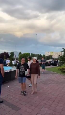 This media file captures a lively scene in Ilidža, Bosnia and Herzegovina, likely during an evening gathering. The setting appears to be a public space, possibly a park or plaza, featuring a decorative fountain with a whimsical, abstract sculpture at its center. The fountain's edge is lined with people, some sitting and conversing, while others stand or walk around.

The subjects are primarily people of various ages, dressed casually for what seems to be a social event. In the foreground, two men are walking. One man, wearing a dark blue t-shirt and blue trousers, is walking away from the camera. Another man, in a black t-shirt and shorts, walks towards the camera. A young girl with a ponytail is seen near the fountain, wearing a white t-shirt and a blue skirt.

In the middle ground, a group of people are seated on the edge of the fountain, enjoying each other's company. A bouquet of flowers rests on the fountain's edge near a couple. Further back, more people are gathered, suggesting a communal event or celebration.

The time of day appears to be late afternoon or early evening, as indicated by the soft, diffused light and the cloudy sky, which has a dramatic, slightly overcast appearance. The presence of streetlights in the background also hints at the approaching evening. The overall mood seems relaxed and social, with people engaging in conversation and enjoying the outdoor space.

Notable details include the unique sculpture on the fountain, which resembles a character with multiple colorful appendages emerging from its head. The ground is paved with reddish-brown bricks in a herringbone pattern. In the background, trees and some distant buildings are visible, along with what appear to be utility poles and possibly cranes, suggesting an urban or semi-urban environment. There is visible text on the t-shirt of one of the men in the foreground: "THE NORTH FACE". Another man in the middle ground is wearing a t-shirt with the word "BARCELONA" and an emblem.
