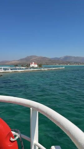 The video shows a view from a boat as it approaches a small island with a white building and an orange roof, possibly a church, situated in turquoise waters under a clear blue sky. In the background, a mountainous landscape is visible. A pier extends from the island towards the boat, with lights lining its edge. The water is slightly choppy, and the railing of the boat is visible in the foreground.