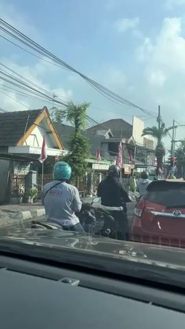 The video shows a street scene from the interior of a vehicle. The foreground consists of the vehicle's dashboard and windshield. Outside, several people on motorcycles are visible, some stopped at what appears to be a traffic light. One person in the middle ground is wearing a light-colored shirt and a blue helmet, and is on a motorcycle. Behind them, a red car is visible. The background shows buildings and trees, with utility lines overhead. The sky is partly cloudy. Several small flags, likely national flags, are displayed on poles attached to a building on the left. The overall impression is of moderate traffic on a sunny day.