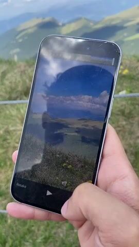 A person is holding a smartphone with a cracked screen in their right hand. The phone displays an image of a mountainous landscape, with text overlaying the image. The background is a grassy hillside with a clear blue sky dotted with white clouds. The phone is being held up to take a photo or video of the scenery. The person's thumb is visible on the bottom of the screen, appearing to interact with the device. The phone's screen reflects the sky and the surrounding landscape.