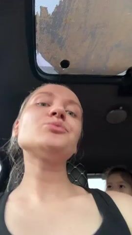 The video shows a woman and a young girl in a car. The woman, who is driving, has fair skin, light eyes, and a nose ring. She is wearing a black tank top. The girl is sitting in the back seat. She is wearing a pink shirt. There is a stuffed cat toy on the seat between them. The camera is positioned in front of the woman, and she is taking a selfie. The girl is smiling and laughing. The car has a sunroof and custom seat covers.