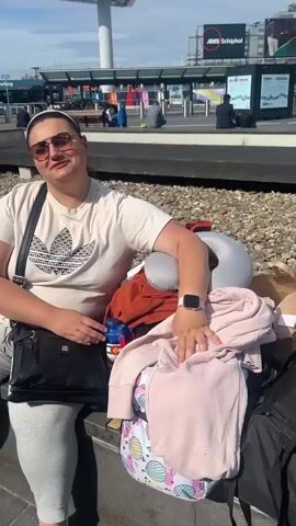 The video shows a woman sitting on a low ledge near a bus stop outdoors. She is wearing sunglasses, a white t-shirt with an Adidas logo, grey sweatpants, and a white headband. She has a black purse slung over her shoulder and is wearing a pink smartwatch.  She reaches into her purse and then gestures with her hands as she speaks. Next to her on the ledge is an orange sweater, a pink jacket, a neck pillow, and a water bottle.  In the background, there is a bus stop with several people and buses. A sign reads "AMS Schiphol Parking." Above the bus stop, there is a large sign with text in white and green. The sky is blue with some clouds.