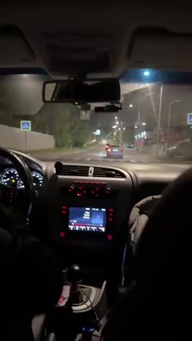 The video shows the interior of a car at night, from the perspective of a passenger looking forward. The dashboard is visible, with a red-lit stereo system and other controls. The driver's hands are visible on the steering wheel on the left side of the frame. The road ahead is illuminated by streetlights and the headlights of other vehicles. A car is seen driving in front of the vehicle. The environment appears to be a street or road lined with trees and buildings.
