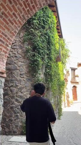 The video shows a person walking down a cobblestone street between buildings with stone and stucco walls covered in greenery. The person filming is wearing a black shirt and a gold chain. They pass a group of people walking in the opposite direction. The buildings are in a rustic, old-world style with tile roofs. One building has a sign with a green arrow pointing to an exit. The sky is blue.