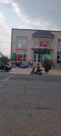 The video shows a two-story building with shops on the ground floor. A sign above one of the shops reads "MAGAZIN ORO". There are people on the sidewalk and a car is parked in front of the building. A person is riding a scooter on the street in front of the building. The weather is cloudy.