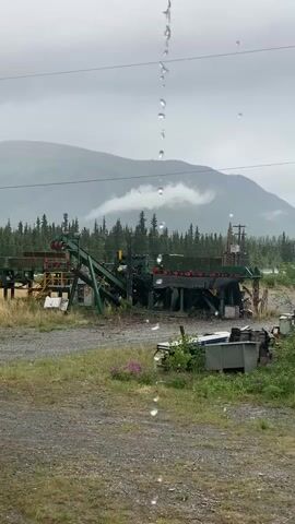 The video shows a conveyor belt system in an outdoor setting under overcast skies, with rain falling in the foreground. The conveyor belt system is green and appears to be industrial or for agricultural use. It is situated in a gravel and grassy area. In the background, there are trees and mountains, with some clouds obscuring the peaks. The overall impression is one of a remote, possibly rural or wilderness location experiencing inclement weather. There are no people or animals visible in the video. The rain is depicted as a series of water droplets falling vertically, creating a foreground element that obscures the view slightly.