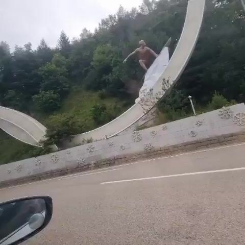 The video shows a sculpture of a man, possibly a statue, positioned on a hillside. The sculpture appears to be in various dynamic poses, one of which resembles a skier in motion, with a white, abstract form underneath that could represent snow or water. The sculpture is located above a curved concrete structure that resembles a bridge or a large arch, with a retaining wall adorned with snowflake-like decorative elements. A road runs in front of the retaining wall, and the video is filmed from the perspective of someone inside a vehicle, as evidenced by the car's side mirror visible in the lower left corner. The environment is natural, with a densely wooded hillside and overcast sky.