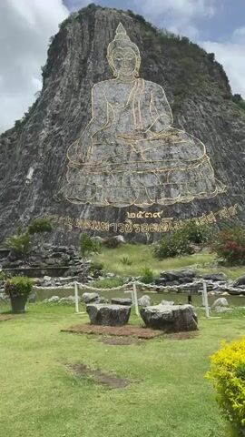 A large, golden Buddha image is carved into a rocky cliff face. The Buddha is depicted in a seated position with hands in a meditative pose. Below the Buddha image, there is text inscribed in Thai script. The cliff is grey and textured, with some greenery growing on its lower slopes. In the foreground, there is a grassy area with several large, grey rocks. Beyond the rocks, there is a body of water and a chain-link fence. The sky above is cloudy. The camera is positioned at a low angle, looking up at the cliff face and the Buddha image.