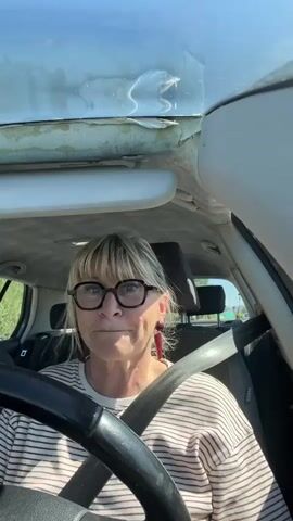 A woman wearing glasses and a striped shirt is driving a car. She is looking directly at the camera with a neutral expression. The car's interior shows wear and tear on the roof. There is music playing. The background appears to be a highway with green vegetation visible.
