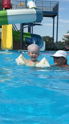A young child wearing a swim cap and arm floaties is splashing in a swimming pool. An adult woman wearing a white hat and sunglasses is standing behind the child. In the background, there is a water slide and other people on lounge chairs. The sky is blue with some clouds. The video appears to be taken on a sunny day.