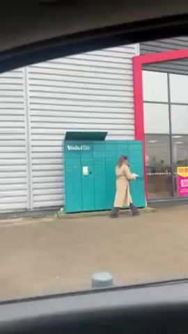 The video shows a woman standing in front of a Vinted Go locker outside a building. She is wearing a tan trench coat and dark boots. She is using the locker, presumably to pick up or drop off a package. The building has a gray wall with horizontal lines and a red frame around its glass front. There is a sign on the window that says "SOLDES 50%." The video is taken from inside a car, looking out the window.
