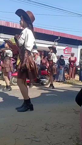 Two young women in cowboy hats and brown ruffled skirts are marching down a paved road. The woman in the foreground is on the left, wearing a white shirt, brown vest, brown skirt, black boots, and black gloves. She is holding something small and yellow in her left hand and looking to her left. The woman behind her, on the right, is also wearing similar attire and appears to be holding a microphone. A red scooter with two people on it is parked on the side of the road behind the marching women. A building with a red awning and a sign that reads "KOPER" is visible in the background. Several other people are standing and watching the parade. The ground is dusty and appears to be a dirt road next to the paved section. The sky is clear and blue.