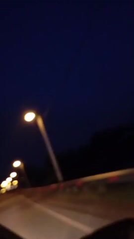 The video shows a nighttime view from inside a vehicle traveling on a highway. Streetlights illuminate the road and the dark, tree-lined landscape bordering it. The camera is positioned to capture the road ahead and to the side, with a partial view of the car's interior in the foreground, including what appears to be a side mirror. The road has multiple lanes, and the guardrail on the right side of the road is visible with reflective markers. The sky is a deep blue, indicative of twilight or early night. The motion blur suggests the vehicle is in transit.