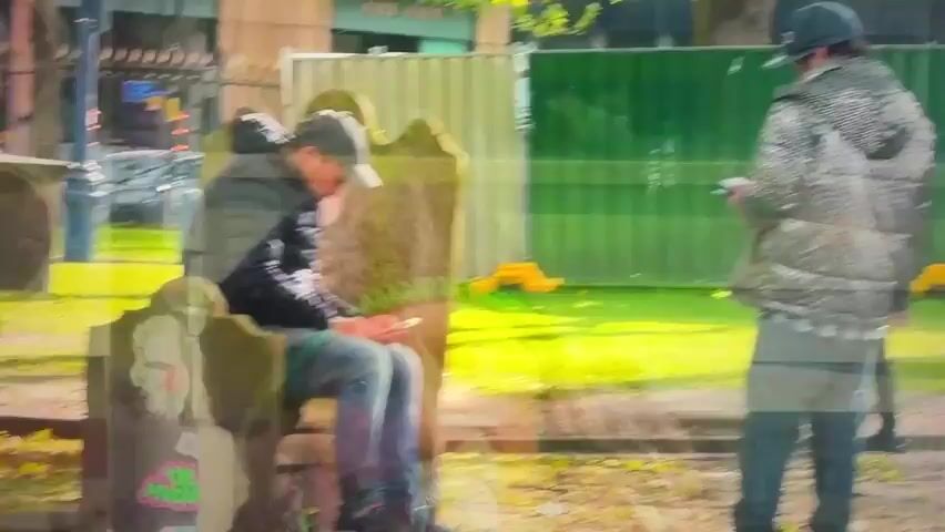 The video shows a public outdoor area, possibly a park or cemetery, with stone structures resembling gravestones or monuments. In the foreground, a man wearing a gray baseball cap, a dark puffer jacket, and blue jeans is sitting on a stone bench, looking down at his phone. To his right, a man in a patterned jacket and blue jeans stands facing away from the camera, also looking at his phone. In the middle ground, two people are walking past, a man in a black jacket and gray pants and a woman in a black jacket and a gray skirt with black tights. Further in the background, a man in a dark suit is walking. The environment appears to be sunny with green grass and trees. There is a green construction fence in the background.