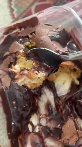 A close-up shot focuses on a spoon lifting a piece of chocolate-covered pastry filled with cream. The pastry is partially submerged in a rich, dark chocolate sauce, with shards of milk chocolate and scattered green pistachios decorating the top. The background shows a patterned fabric, possibly a tablecloth. The lighting is somewhat dim, highlighting the glossy texture of the chocolate.