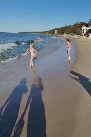 This video captures a sunny day at the beach in Pionersky, Russia. The scene is dominated by a wide, sandy beach with gentle waves lapping at the shore. The sky is a clear, vibrant blue, indicating fair weather and a pleasant time for outdoor activities. The time of day appears to be late afternoon or early evening, as evidenced by the long shadows cast by the people on the beach and the warm, golden light.

The primary subjects are two young girls playing near the water's edge. They are wearing swimsuits, suggesting they are enjoying the beach and possibly swimming or wading. One girl is bending down, possibly interacting with something in the sand or water, while the other is running along the shoreline. Their playful energy and engagement with their surroundings convey a sense of joy and freedom.

In the background, other beachgoers can be seen scattered along the coastline, some swimming in the sea and others relaxing on the sand. Further back, a line of trees and buildings suggests a town or resort area bordering the beach, common for a coastal location. The presence of people and the well-maintained beach point to this being a popular recreational spot.

The video is shot from a low angle, likely by someone walking or standing close to the girls. This perspective emphasizes the vastness of the sea and sky and places the viewer directly into the experience of being on the beach. The long, elongated shadows in the foreground add a dynamic visual element and reinforce the time of day. The overall mood is one of relaxed enjoyment and the simple pleasures of a summer day by the sea. No visible text is present in the media.