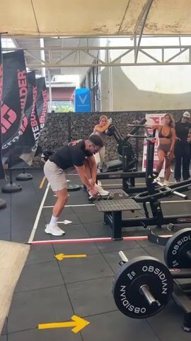 The video shows a man in athletic wear demonstrating a leg exercise on a piece of gym equipment. He is wearing a black t-shirt, light-colored shorts, and white sneakers. He is positioned on a platform with his left foot on a movable footplate. He is reaching forward with his right hand to hold onto a support bar. He appears to be in the process of lifting his left leg. Behind him, several other people are visible, including women in athletic attire, who seem to be observing or participating in the exercise. The environment is a gym with black rubber flooring, various exercise machines, and banners with the text "GLUTEBUILDER". There are also weight plates visible in the foreground. The lighting suggests an outdoor or semi-outdoor setting with overhead structures.