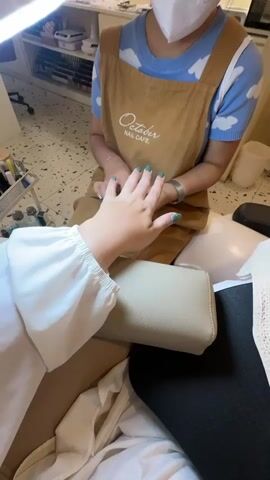 The video shows a person's hand receiving a manicure. A manicurist, wearing a light blue shirt with white cloud patterns and a brown apron with "October Nail Cafe" printed on it, is seen applying nail polish to the fingernails of the person whose hand is in the foreground. The person receiving the manicure is wearing a white top with puffed sleeves. The environment appears to be a nail salon, with shelves containing nail polish bottles and other beauty supplies visible in the background. The lighting is bright, likely from a nail lamp. The camera angle is from the perspective of the person getting their nails done.