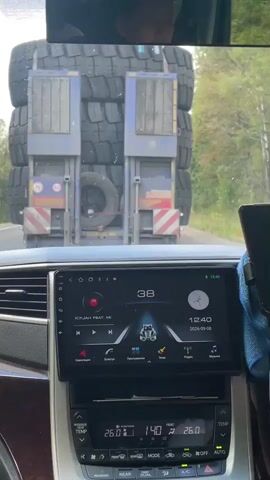 The video shows the interior of a car, viewed from the driver's perspective. The car is on a road, following a large truck loaded with tires. The truck's back is visible, with its taillights and reflective markings clearly seen. The car's dashboard is also in focus, showing a touchscreen infotainment system displaying music information and a clock. The temperature inside the car is displayed as 26 degrees Celsius. A person's face is reflected in the rear-view mirror, suggesting the driver is male. The surrounding environment appears to be a rural or suburban area with trees visible on the side of the road. The video is shot during daylight.