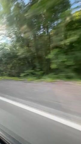This media file captures a view from a moving vehicle, likely a car, traveling along a road. The scene is dominated by a blur of green trees and foliage on the right side, indicating a roadside lined with vegetation. The road itself is visible in the foreground, with a distinct white line marking the lane. A black side mirror of the vehicle is partially visible in the lower left corner. The sky above is blue with scattered white clouds. The overall impression is one of a journey through a natural, possibly rural, landscape on a bright day. The motion blur suggests the vehicle is in transit, and the activity depicted is simply traveling. The time of day appears to be daytime, given the clear sky and sunlight. There are no people clearly visible in the frame. A yellow sign is partially visible on the left side of the road in the distance, but its text is illegible due to the motion and distance. The location context provided is Кулмас, Russia, which aligns with a rural or natural setting. The emotions conveyed are neutral, focusing on the experience of travel.