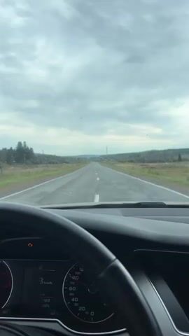 The video shows a view from inside a car as it drives down a two-lane road. The steering wheel and part of the dashboard are visible in the foreground. The road stretches ahead into the distance, with fields and trees on either side. The sky is cloudy.