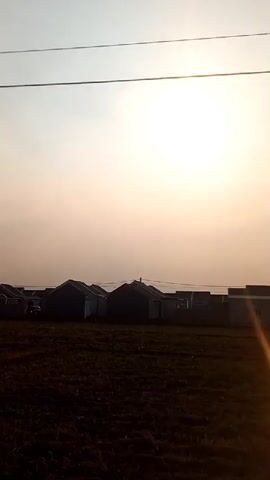 The video shows a row of houses with light-colored walls and dark roofs. In front of the houses is a field of dry, brown grass. The sun is shining brightly in the sky, which is a pale yellow and white, suggesting it is either sunrise or sunset. There are power lines visible in the sky above the houses. The camera appears to be moving, as the perspective of the houses and field changes slightly throughout the short clip. There are no people or vehicles visible in the frame.