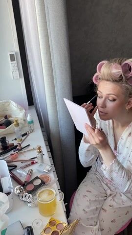 A woman sits at a table near a window, applying makeup. She wears pink and white floral patterned pajamas and has pink hair rollers in her blonde hair. The table is covered with various makeup items including brushes, bottles, and palettes. There is a mug of yellow liquid, possibly beer or juice, near the makeup. She holds a small mirror in her left hand and uses a makeup brush in her right hand to apply makeup to her face. The window behind her is covered with gray curtains. A white remote control is mounted on the wall near the window.