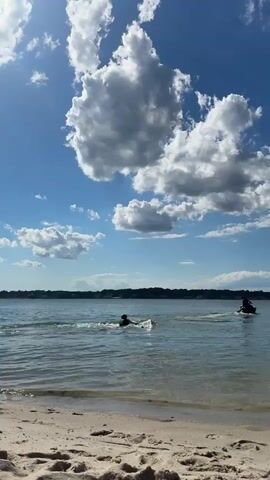 The video shows a body of water with a sandy beach in the foreground. Two people are on jet skis in the water. The person on the left jet ski is standing up and appears to be in motion, leaving a wake behind them. The person on the right jet ski is also in motion and leaving a wake. The sky is blue with large, white clouds. There is a distant shoreline with trees.