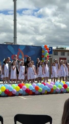 A children's choir is performing outdoors on a stage.  The choir is comprised of approximately 15 young girls, who are wearing white dresses with butterfly appliques. They are accompanied by several adult women, two of whom are holding microphones.  A colorful balloon arch adorns the front of the stage.  The stage backdrop is a blue banner with an abstract design. A parking sign is visible next to the stage.  A building can be seen in the background. The sky is cloudy. An audience can be partially seen in the foreground.