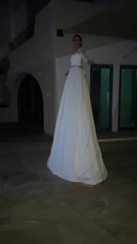 The video shows a woman in a white, floor-length dress standing in an outdoor space at night. The dress appears to be a wedding dress, with a fitted bodice and a flowing skirt. She is wearing a belt around her waist. Her hair is pulled back. In the background, there is a white building with arched doorways and windows. The ground is a dark, textured surface. The lighting is dim. The woman is facing away from the camera, then turns to look at the camera briefly.