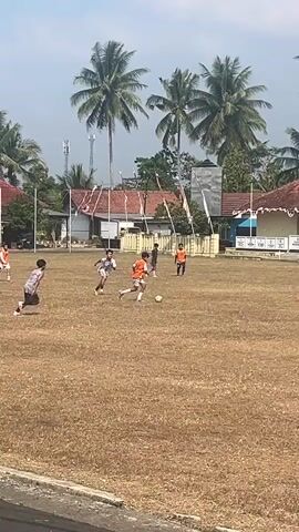 A soccer game is being played on a dry, brown field. There are several young people, appearing to be teenage boys, on the field. They are wearing various colored jerseys and shorts. Some are running, some are kicking a soccer ball, and some are standing. In the background, there are palm trees, buildings with red tile roofs and white walls, and some flags. The sky is a light blue with some white clouds. The ground is covered in dry grass. The overall scene appears to be an outdoor sporting event in a tropical or subtropical climate.