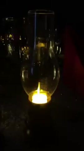 The video shows a small, clear glass lantern with a tea light candle inside. The lantern sits on a dark surface that may be a table. Other glasses and a red napkin are visible in the background, suggesting a dinner setting. The candle flame is lit, providing the only light source. The scene appears to be dimly lit.