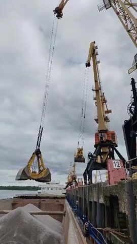 The video shows a harbor with large industrial cranes offloading cargo from a ship. The sky is overcast, and the cranes are visible along the pier. One crane in particular is actively lifting and moving a large clamshell bucket filled with what appears to be gravel or another type of bulk material from the hold of the ship. The number 12 is visible on the side of the crane's base. A white ship can be seen in the background, positioned parallel to the pier.
