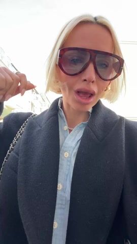The video shows a blonde woman with short hair, wearing oversized sunglasses, a denim button-down shirt, and a black coat. She is outdoors, seemingly on a city street, with buildings and a partly cloudy sky in the background. Initially, she adjusts her sunglasses, then turns to her right, looking around the environment. She takes out a black pug, which she kisses, and then the video ends.