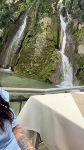 A woman with dark hair and sunglasses is sitting at a table with a white tablecloth, with a waterfall in the background.