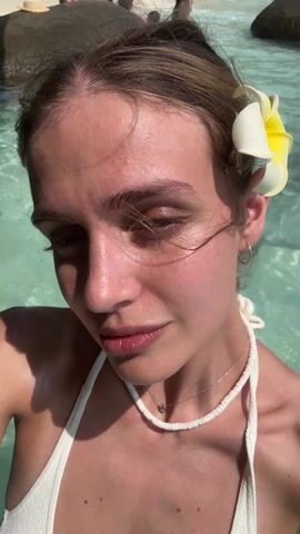 A young woman stands chest-deep in clear, turquoise water. She's wearing a white bikini top, a beaded necklace and a delicate silver chain. A white and yellow plumeria flower adorns her hair. She opens her eyes slightly, then rests her cheek on her left hand, revealing a small tattoo on her wrist. Other people are visible in the background, both in and out of the water.