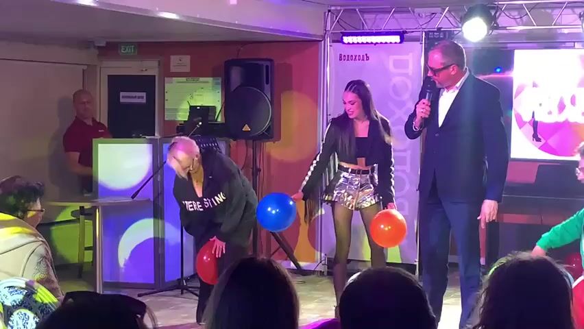 The video shows a stage performance with several people. In the foreground, three individuals are on stage. To the left, a person with short blonde hair, wearing glasses and a black hooded sweatshirt with "WERE STING" printed on it, is crouched down, holding two balloons, one red and one blue. Next to them, a young woman with long dark hair, wearing a black cropped jacket with a silver sequined skirt, is standing and holding a red balloon. To the right, a man in a black suit, wearing glasses, is holding a microphone and appears to be addressing the audience. A DJ booth with speakers and colorful lighting is visible in the background. There are also people seated in the audience in the foreground. The overall environment suggests a live event or entertainment show.
