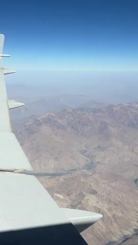 The video shows an aerial view from an airplane window. A white airplane wing is visible in the foreground, taking up the left side of the frame. The background displays a vast, arid mountain range under a clear blue sky. The mountains are rugged and appear to be brown or tan in color, with visible valleys and geological formations. There is a slight haze obscuring the distant mountains. The sun appears to be shining, casting shadows. No people are visible in the video.