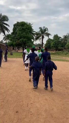 A group of uniformed children march across a dirt field lined with palm trees on a cloudy day. The children are in lines and columns, some carrying flags. One uniformed man walks along the side of the marching children. In the background are mountains and huts.