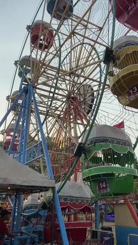 A low-angle shot captures a Ferris wheel with multiple colorful gondolas as it rotates, illuminated by the soft light of what appears to be late afternoon or early morning. The gondolas, some numbered, are predominantly red, blue, and green, with decorative trim. The Ferris wheel's metal structure is a mix of blue and orange. In the foreground, a glimpse of people and what seems to be part of a stall or food vendor can be seen, suggesting an amusement park or fair setting. The sky is a pale, hazy blue, with hints of sunlight filtering through the Ferris wheel's spokes.
