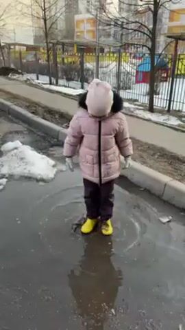 A young child wearing a light pink hooded jacket, dark pants, white gloves, and yellow rubber boots is jumping in a puddle. The child is facing the camera. The puddle is on an asphalt surface next to a curb, with a pile of dirty snow on the other side of the curb. In the background, there's a fence enclosing a playground, and apartment buildings are visible further away. The day appears to be overcast.