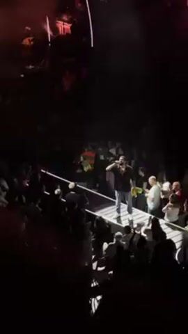 A male singer stands on a white platform in front of a large crowd. He is wearing jeans and a dark shirt. The stage is brightly lit, but the crowd is mostly in shadow. The band is playing behind him on another stage above. He is raising a green towel to the crowd, and the crowd is cheering.